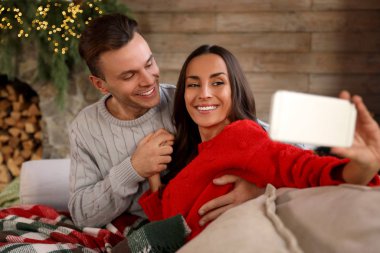 Mutlu genç çift Noel için dekore edilmiş oturma odasında selfie çekiyor.