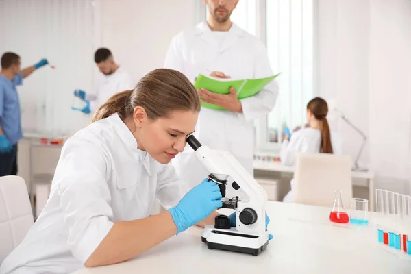 Científico usando microscopio en la mesa y colegas en el laboratorio — Foto de Stock