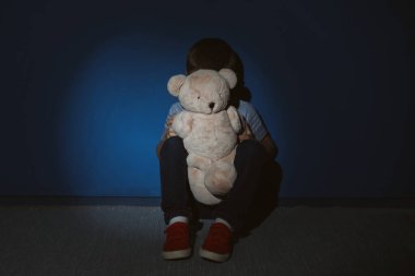 Mavi duvarın yanında oyuncak ayısı olan küçük bir çocuk. Aile içi şiddet suçlusu