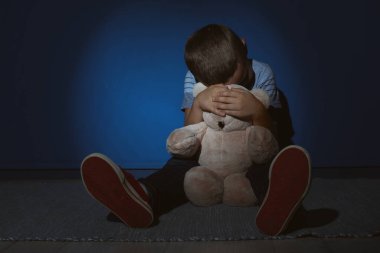 Sad little boy with teddy bear near blue wall. Domestic violence clipart