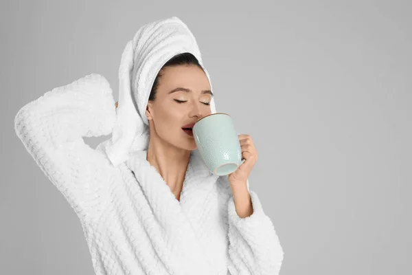 Beautiful young woman in bathrobe with cup of coffee on light gr. — стоковое фото