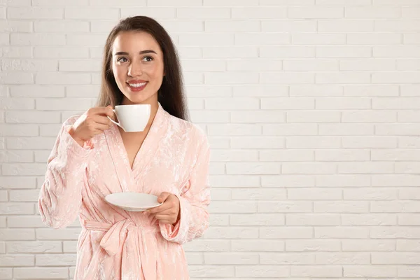 Jovem mulher em roupão de banho com xícara de café perto da parede de tijolo branco — Fotografia de Stock