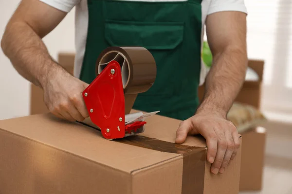 Man packing box with adhesive tape indoors, closeup. Moving serv