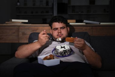 Depressed overweight man eating sweets in living room at night clipart