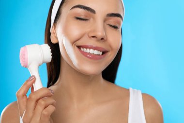 Açık mavi arka planda yüz temizleme fırçası kullanan genç bir kadın, yakın plan. Yıkama aksesuarı