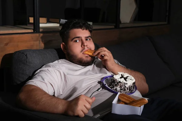 Depressed overweight man eating sweets in living room at night