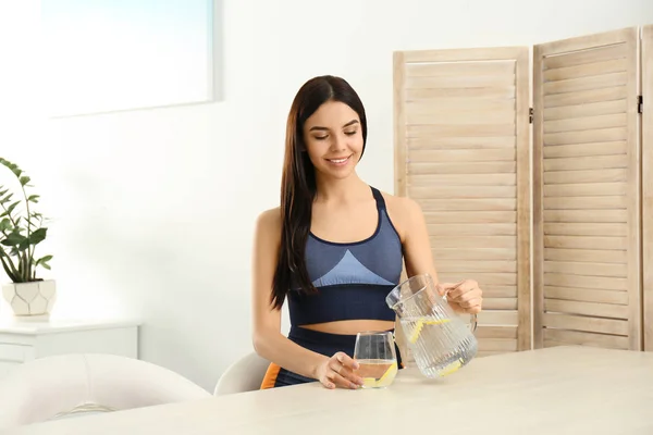 Beautiful young woman pouring lemon water into glass from jug at — ストック写真