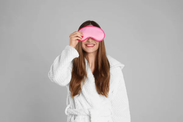 Νεαρή γυναίκα με μπουρνούζι και μάσκα ύπνου στο ανοιχτό γκρι πίσω μέρος — Φωτογραφία Αρχείου