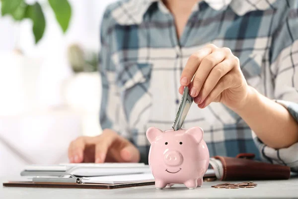 Frau legt Geld in Sparschwein bei Tisch drinnen, Nahaufnahme — Stockfoto