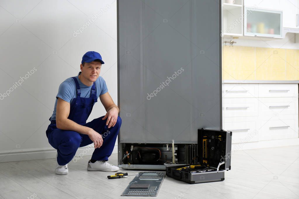 Male technician in uniform near broken refrigerator indoors