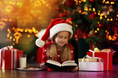 Noel Baba şapkalı küçük kız Noel ağacının yanında kitap okuyor. 