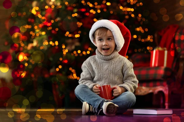 Petit garçon dans la casquette du Père Noël avec boisson près de l'arbre de Noël à — Photo