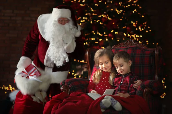 Santa Claus sneaking in with gifts while children reading book n — Stock Photo, Image