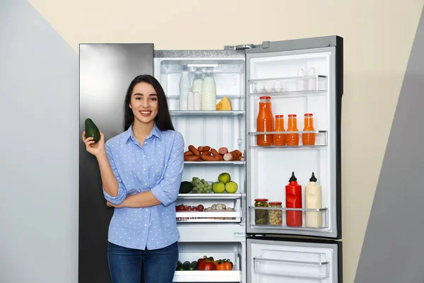 Šťastná mladá žena s avokádem v blízkosti otevřené chladničky uvnitř — Stock fotografie