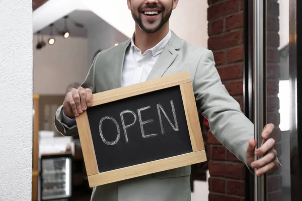 Young business owner holding sign OPEN at door of his cafe, clos