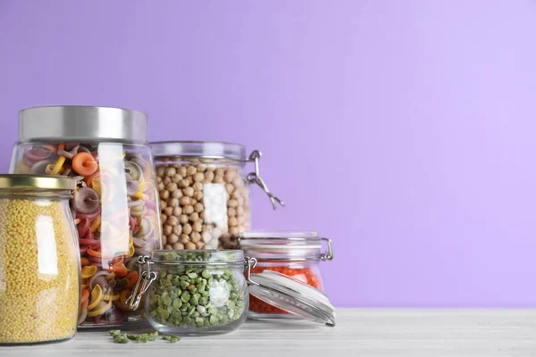 Frascos con diferentes cereales sobre mesa de madera blanca contra violeta — Foto de Stock