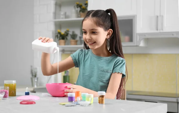 Милая маленькая девочка, наливающая клей в миску за столом на кухне. DIY — стоковое фото
