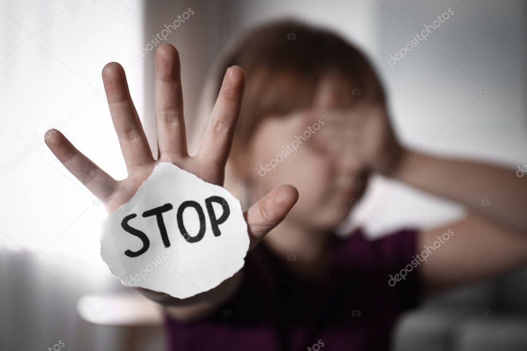 Abused little girl with sign STOP indoors, focus on hand. Domest