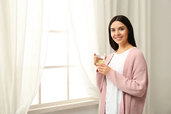 Piękna młoda kobieta ze smaczną wodą cytrynową w pobliżu okna — Zdjęcie stockowe