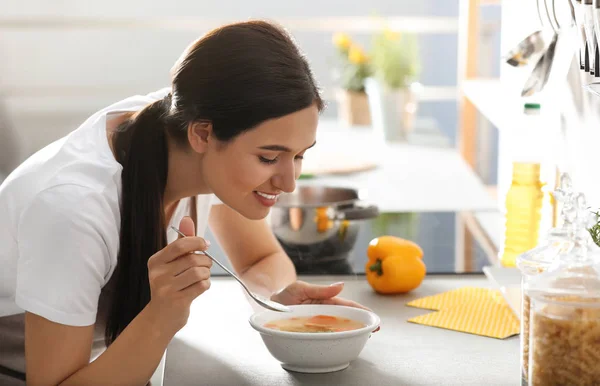 Ung kvinde spiser velsmagende grøntsagssuppe ved køkkenbordet i køkkenet - Stock-foto