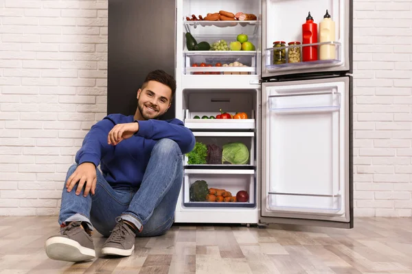 Joyeux jeune homme assis sur le sol près d'un réfrigérateur ouvert à l'intérieur — Photo