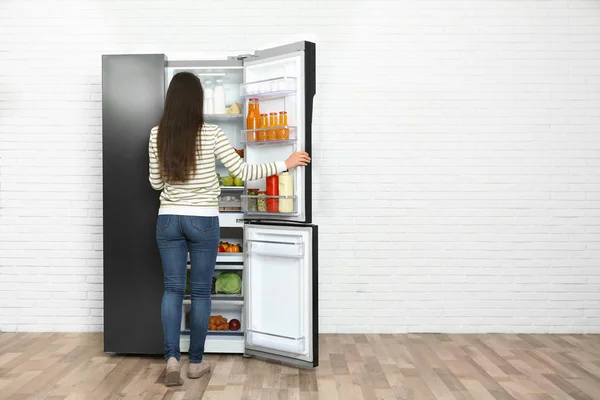 Молодая женщина выбирает еду из холодильника рядом с белым кирпичным валом — стоковое фото