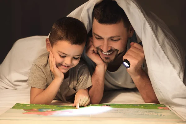 Pai e filho com lanterna livro de leitura sob cobertor em hom — Fotografia de Stock