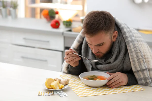 Больной молодой человек ест вкусный суп, чтобы вылечить грипп за столом на кухне — стоковое фото