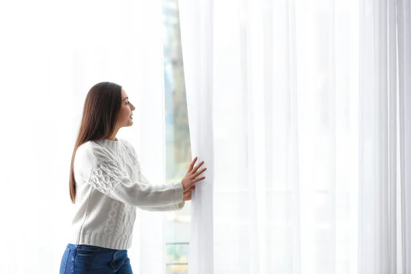 Mujer joven abriendo cortinas de ventana en casa — Foto de Stock