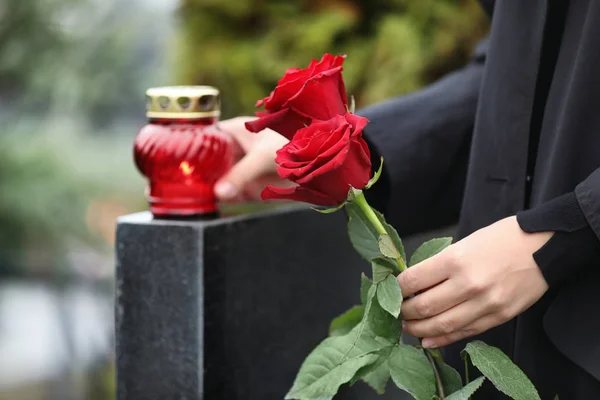 Kobieta trzyma czerwone róże w pobliżu czarnego granitu nagrobek ze świecą — Zdjęcie stockowe