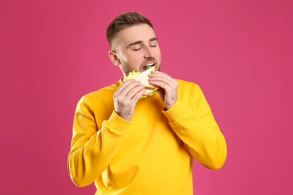 Young man eating tasty sandwich on pink background — Stock Photo, Image