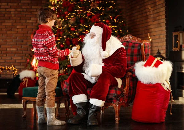 Niño tratando a Papá Noel con galletas cerca del árbol de Navidad — Foto de Stock