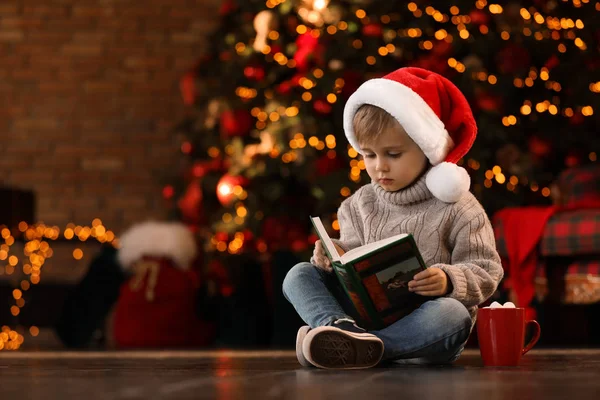 Petit garçon dans le bouchon du Père Noël livre de lecture près de l'arbre de Noël un — Photo