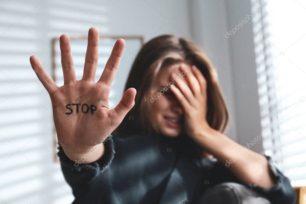 Crying young woman showing palm with word STOP indoors, focus on