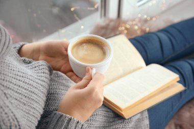 Elinde kahve fincanı olan bir kadın evde kitap okuyor.
