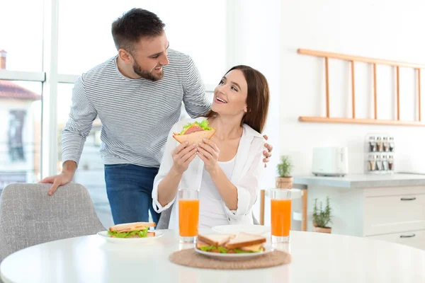 Pareja feliz desayunando con sándwiches en la mesa en kitche —  Fotos de Stock
