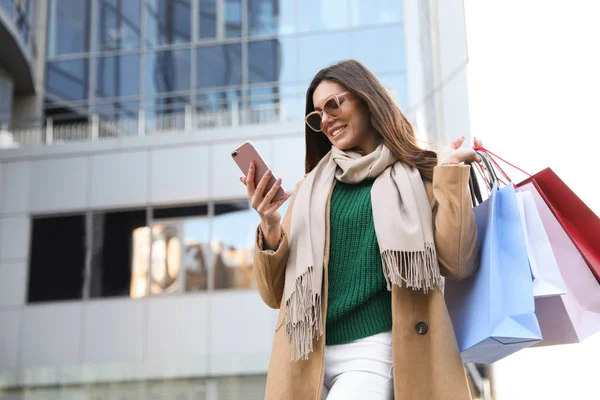 Mooie jonge vrouw met boodschappentassen en smartphone buiten — Stockfoto