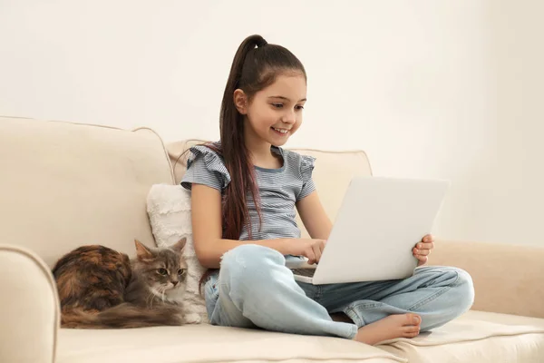 Ragazzina carina con computer portatile e gatto sul divano a casa. Primo animale domestico — Foto Stock