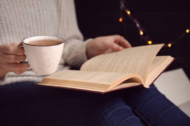 Elinde kahve fincanı olan bir kadın evde kitap okuyor.