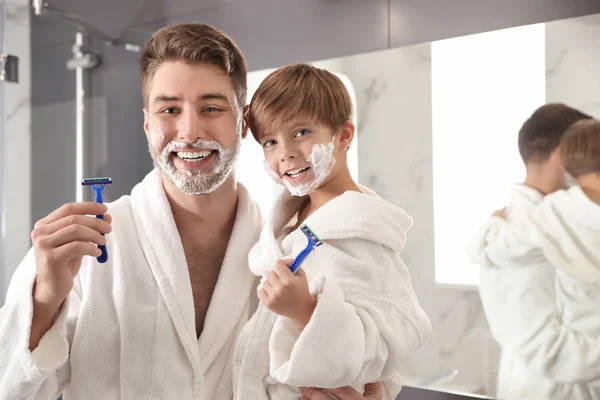 Tata i syn z pianką do golenia na twarzach trzymających brzytwy w szlafroku — Zdjęcie stockowe