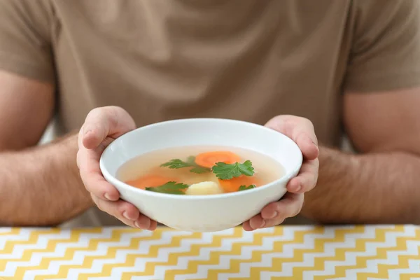 Молодой человек держит миску вкусного супа за столом, крупным планом — стоковое фото