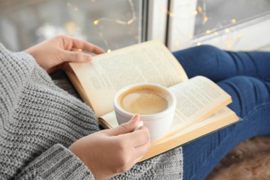 Elinde kahve fincanı olan bir kadın evde kitap okuyor.