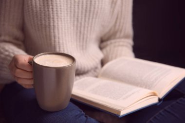 Elinde kahve fincanı olan bir kadın evde kitap okuyor.