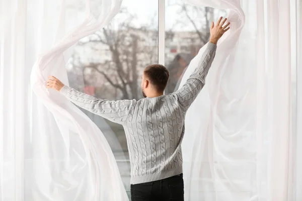 Hombre joven abriendo cortinas de ventana en casa, vista trasera — Foto de Stock