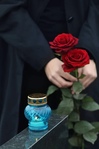 Женщина, держащая красные розы возле черного гранитного надгробия со свечой — стоковое фото