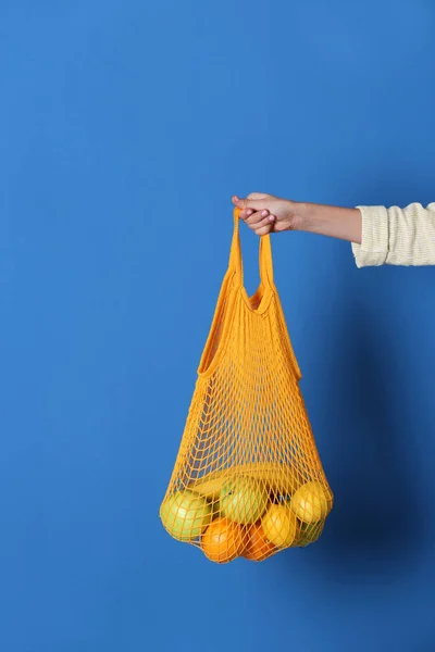 Woman holding net bag with fruits on blue background, closeup