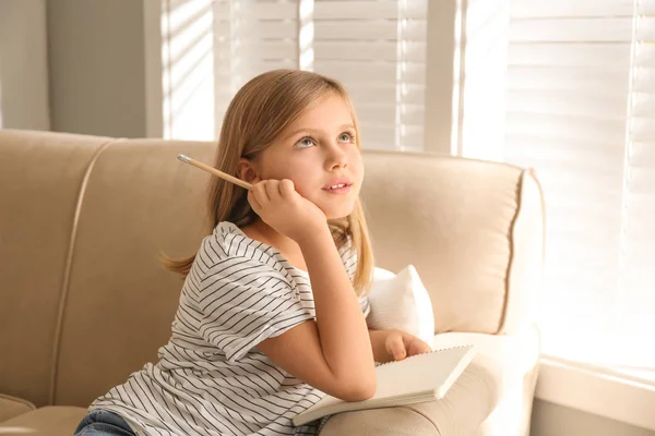 Задумчивая маленькая девочка с карандашом и блокнотом на диване дома — стоковое фото
