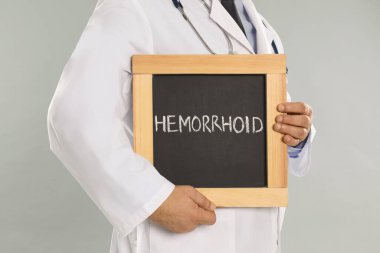 Açık gri arka planda hemoroit sözcüğüyle karatahta tutan doktor, yakın plan.
