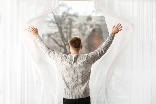 Young man opening window curtains at home, back view — 스톡 사진