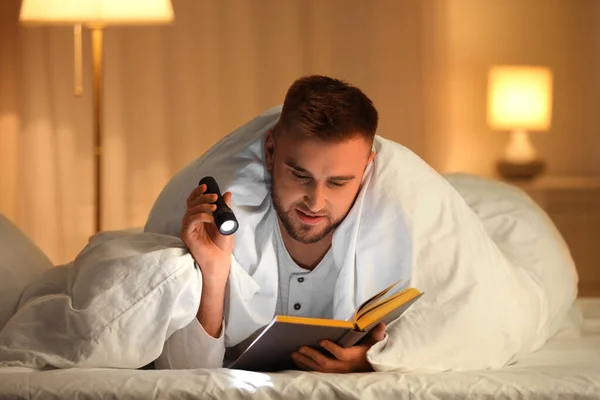 Молодой человек с фонариком читает книгу в спальне — стоковое фото